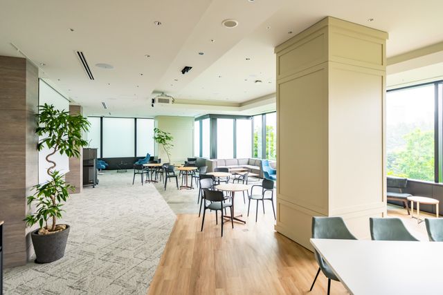 Round cafe tables and dark chairs lead toward lounge seating beside broad windows and indoor plants.