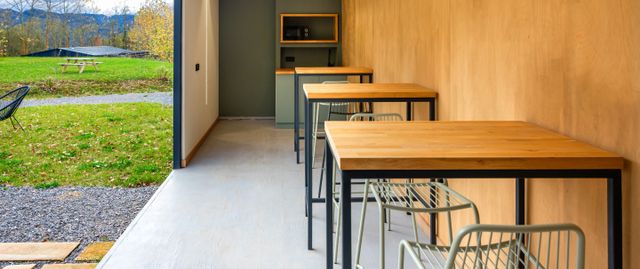 High timber tables and pale metal stools line a wood-paneled wall beside a compact kitchenette and an opening to a garden.