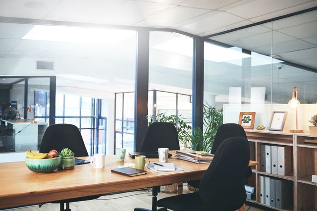 Black chairs surround a long wood-tone table with mugs and notebooks beside open shelving and glass office partitions.