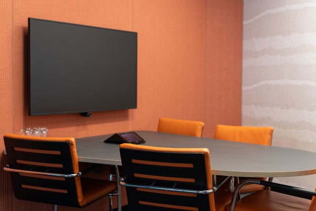 Orange-and-black chairs surround a gray meeting table beneath a wall-mounted screen, with a small tabletop control display.