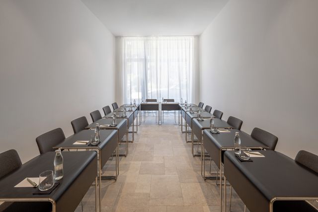 Dark tables and gray chairs form a U-shaped layout in a narrow white room, with water bottles and notepads at the seats.