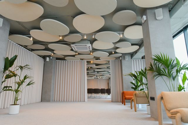 Circular ceiling panels hang above lounge chairs and potted plants, with curtains opening onto rows of chairs in a meeting area.