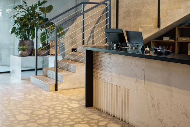 A pale textured reception counter with a dark top stands beside a cable-railed staircase and large potted plants.