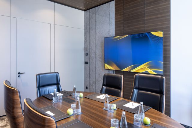 Black high-backed chairs surround a wood-tone meeting table set with notepads and water bottles beneath a wall-mounted screen.