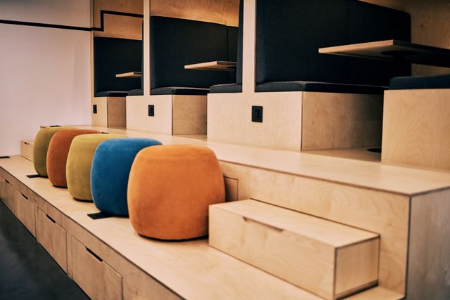 Dark upholstered booths with small tables sit on a raised wood-tone platform above orange, yellow and blue stools.