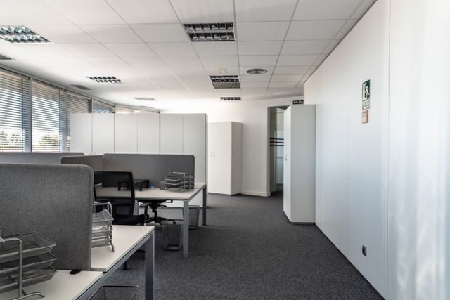 White desks with gray divider panels and black task chairs sit beside window blinds, with storage cabinets along the back wall.