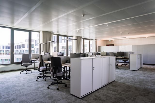 Desks with monitors, gray divider panels and task chairs sit beside tall windows, with white storage cabinets between desk groups.