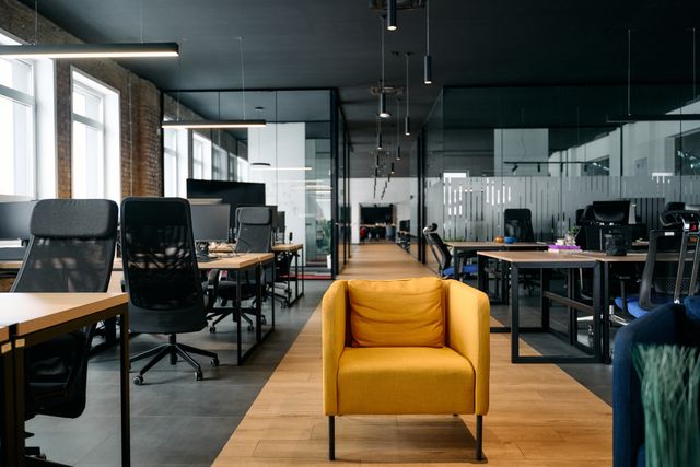 A yellow armchair stands between rows of desks and black task chairs, with glass-partitioned rooms and suspended lights behind.