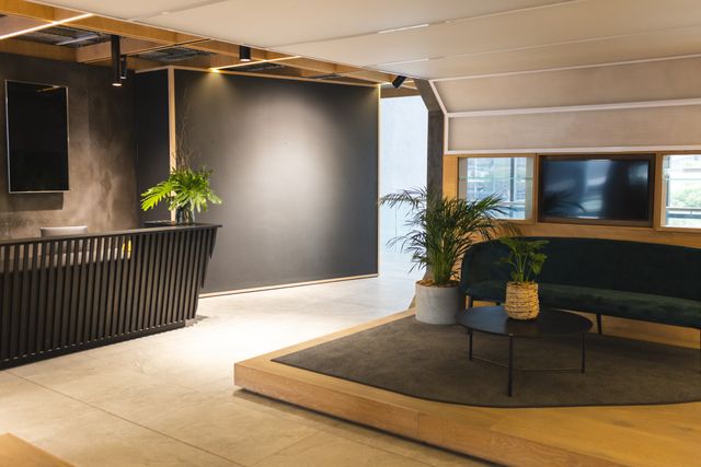 A slatted dark reception counter stands beside a raised seating area with a dark green sofa, round table and potted plants.