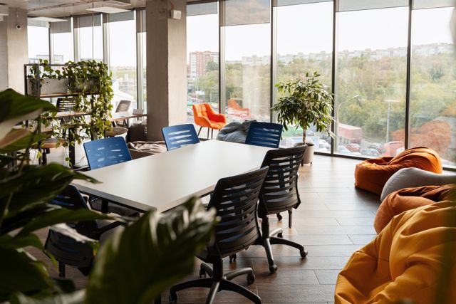 Blue and black task chairs surround a white table beside colorful beanbags, leafy plants and floor-to-ceiling windows.