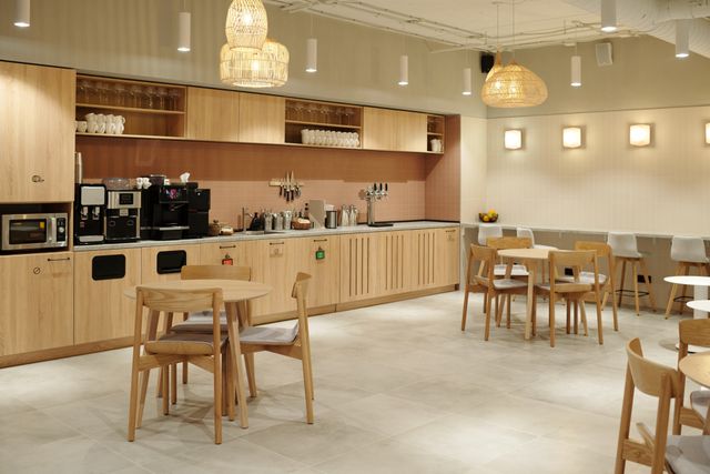 Timber cabinetry, coffee machines and a pink-tiled backsplash line a shared pantry with round tables, chairs and woven pendant lights.