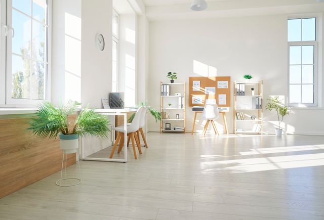 Two white desks and chairs sit between tall windows in a bright room with open shelves, a pinboard and potted plants.