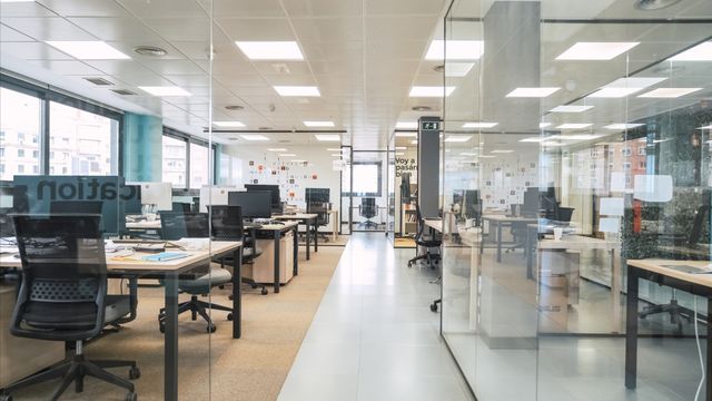 Rows of desks, monitors and black task chairs sit beside glazed partitions and a central aisle in a bright office.