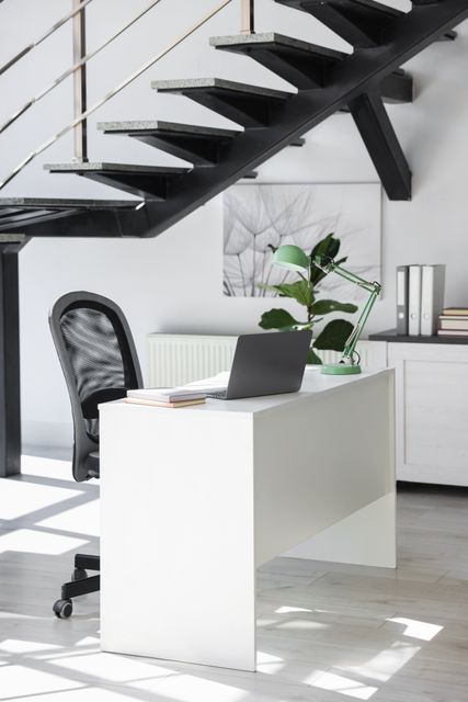 A white desk with a laptop and green task lamp sits beneath an open staircase, alongside a black mesh chair and storage cabinet.