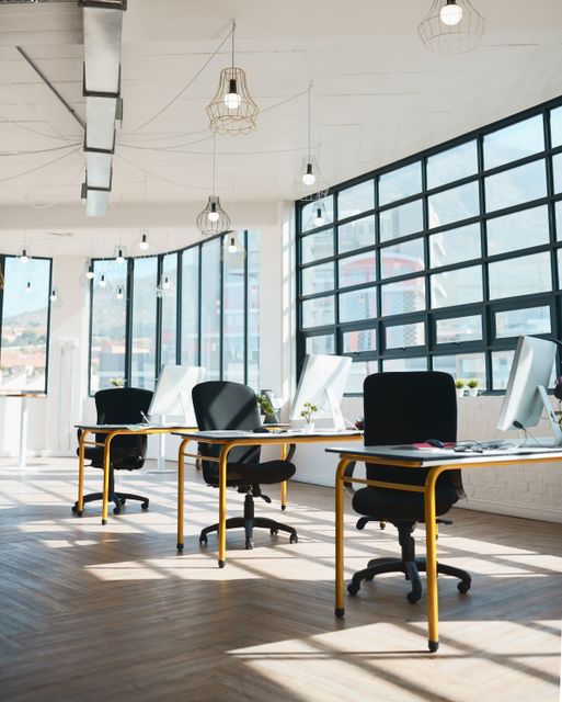 Desks with yellow legs, white computer monitors and black task chairs line a large window wall beneath wire-frame pendant lights.