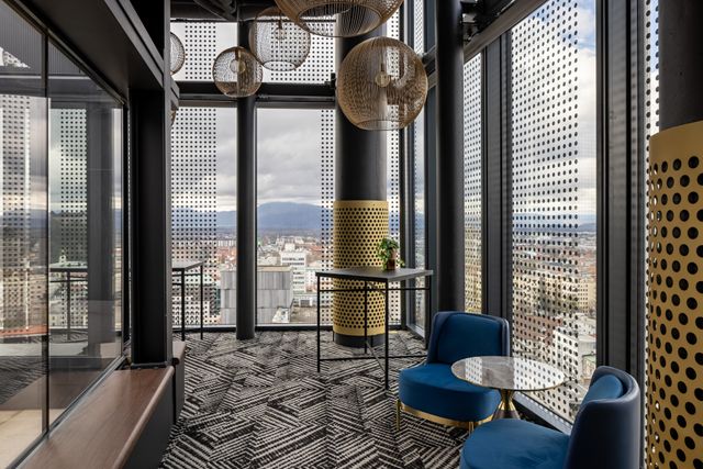 Blue lounge chairs and a round table sit beside tall dot-patterned windows overlooking city buildings and distant hills.