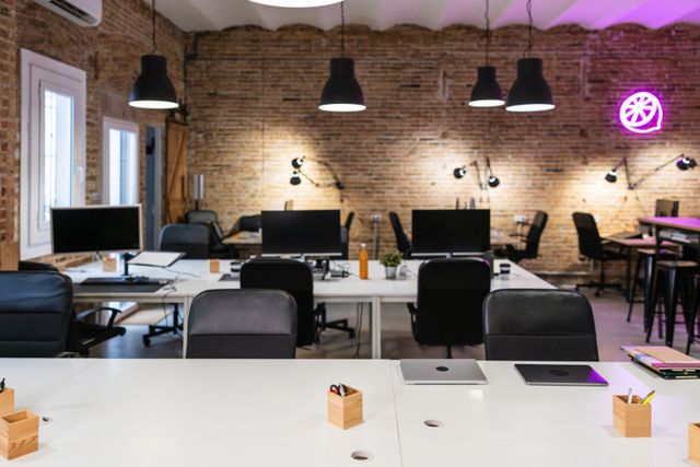 White shared desks, computer monitors and black chairs fill a room with brick walls, black pendant lights and a purple illuminated symbol.