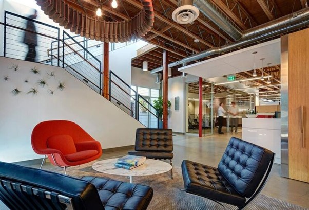 The image showcases an industrial-style office lobby and waiting area featuring black tufted leather chairs, a vibrant red accent armchair, exposed wooden ceiling beams, and a prominent staircase.