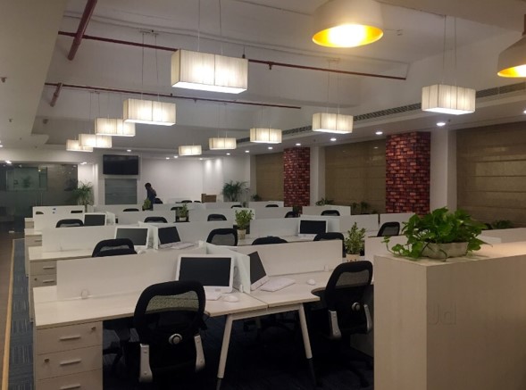 This shows a spacious, professional office floor plan featuring rows of white cubicle desks equipped with computers, black rolling office chairs, warm overhead pendant lighting, and brick-patterned accent pillars.