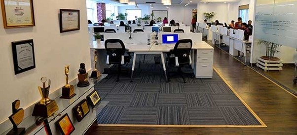 This image depicts an open-plan corporate office with rows of white desks and mesh chairs, framed by a prominent display of awards and certificates on the foreground left wall.