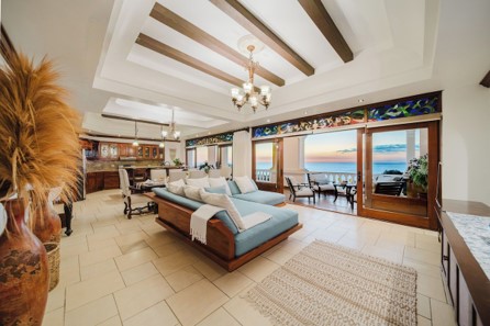 This image showcases a luxurious oceanfront penthouse living room featuring a recessed tray ceiling with wooden beams, custom wooden furniture with light blue cushions, and large sliding doors that open to a scenic sunset sea view.