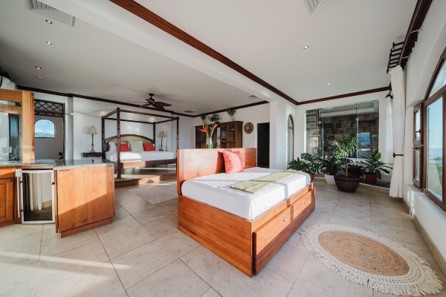 This image shows an expansive, open-concept tropical suite featuring two warm wood beds—including a four-poster frame—complemented by large floor tiles, potted indoor plants, and a small built-in bar area.
