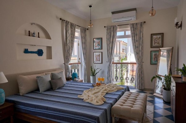 The image showcases a charming Mediterranean-style bedroom with a blue striped bedspread, checkered floor tiles, arched wall niches, and French doors that open up to a sunny balcony view.