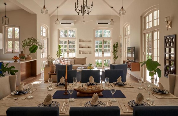 This image illustrates an elegant, open-concept villa interior featuring a meticulously set formal dining table that transitions into a sunlit living space with arched French doors, high ceilings, and a classic central chandelier.